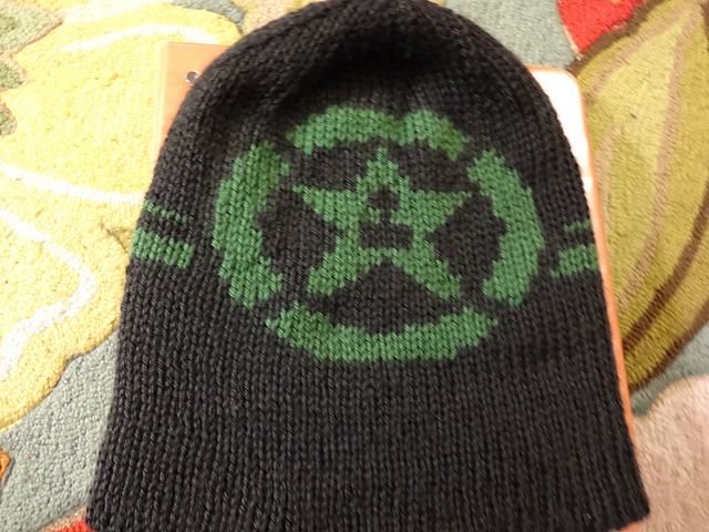 A tall, thick, handknit black beanie with the Achievement Hunter logo in green. The logo depicts the xbox achievement icon, but with a star that has a joystick cutout in place of the trophy.