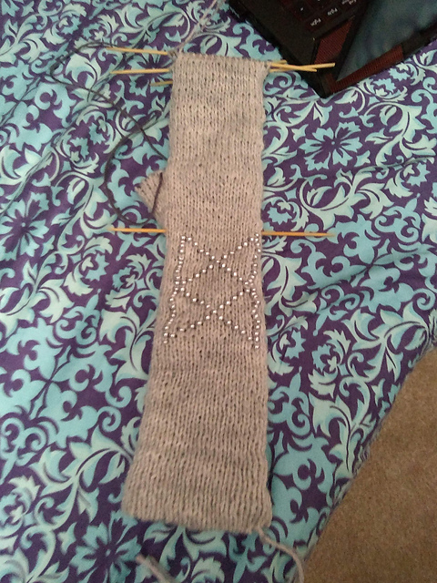A grey, handknit fingerless glove with silver beads making the symbol of the Bureau of Balance from The Adventure Zone.