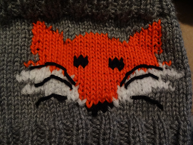 A grey handknit object with an orange and white fox stitched over it. Most of the fox is in a pixel art style matching the shape of the stitches, but the whiskers are embroidered on top with no regard for the grid.