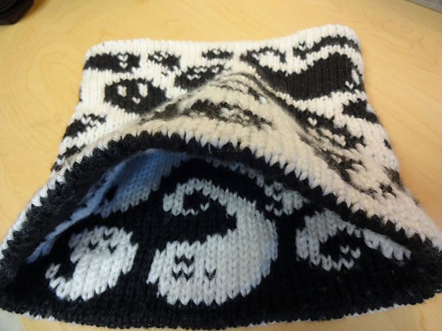 A handknit cowl showing many whimsical black ghosts on a white background around the outside, with one edge propped up to show a peek of the reverse-color mirror image on the inside.