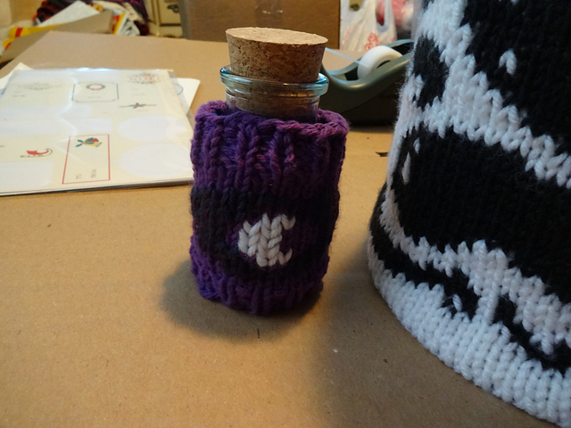 A small jar with a cork stopper, swaddled in a purple handknit cozy that depicts the moon-iris eye logo of the podcast Welcome to Night Vale.