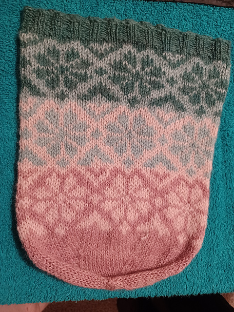 A tall, knit, 4-colored beanie laid flat on the table. The crown of the hat is a dark pink, and the short ribbed brim is a dark green. Using three bands of fair isle flowers with some geometric flourishes, the hat transitions from dark pink to light pink to light green to dark green.